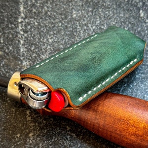May include: A green leather lighter case with white stitching, holding a silver and red lighter. The case is lying on a gray textured surface.
