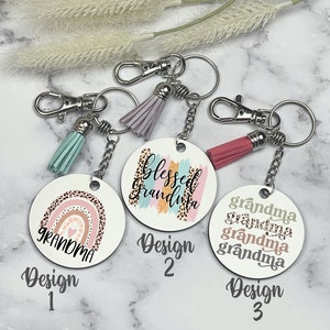 May include: Three keychains with different designs. The first keychain has a rainbow with the word "grandma" on a white background. The second keychain has a white background with the words "blessed grandma" in pink, blue, and gold. The third keychain has a white background with the words "grandma" in different shades of pink, green, and brown.