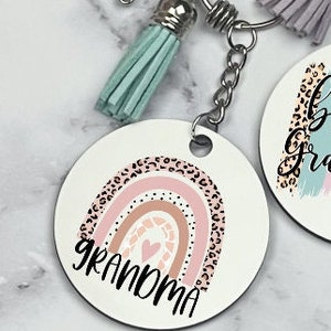 May include: A white round keychain with a rainbow design and the word "grandma" in black lettering. The rainbow has a pink, brown, and beige color scheme with a black and white leopard print pattern. The keychain is attached to a silver key ring with a turquoise tassel.
