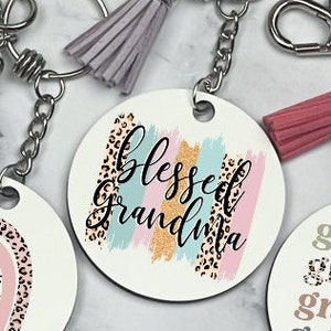 May include: A white round keychain with a pink, blue, and gold brushstroke design and a leopard print pattern. The text "blessed grandma" is written in black cursive font.
