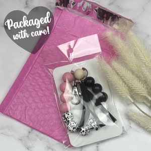 May include: A pink, black, and white beaded keychain with a cow print charm. The keychain is packaged in a pink bubble mailer with the text "Packaged with care!" on it.