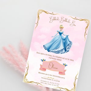Puede incluir: Una invitación rosa y dorada con una imagen de dibujos animados de Cenicienta con un vestido azul. La invitación dice "Bibbidi-Bobbidi-Boo" y "Érase una vez un cumpleaños, en un reino no tan lejos, una princesa llamada Olivia te da la bienvenida a su fiesta de cumpleaños número 2 Sábado 25 de diciembre, a las 8:30 PM 123 Anywhere St., Any City, ST 12345 RSVP a Mark, Teléfono: 123-450-7880"