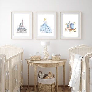 May include: A nursery room with two white cribs, a wooden table with a lamp, and three framed prints on the wall. The prints feature a watercolor illustration of Cinderella's castle, Cinderella in her blue gown, and Cinderella's carriage.