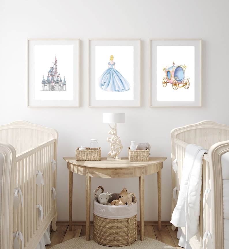 May include: A nursery room with two white cribs, a wooden table with a lamp, and three framed prints on the wall. The prints feature a watercolor illustration of Cinderella's castle, Cinderella in her blue gown, and Cinderella's carriage.