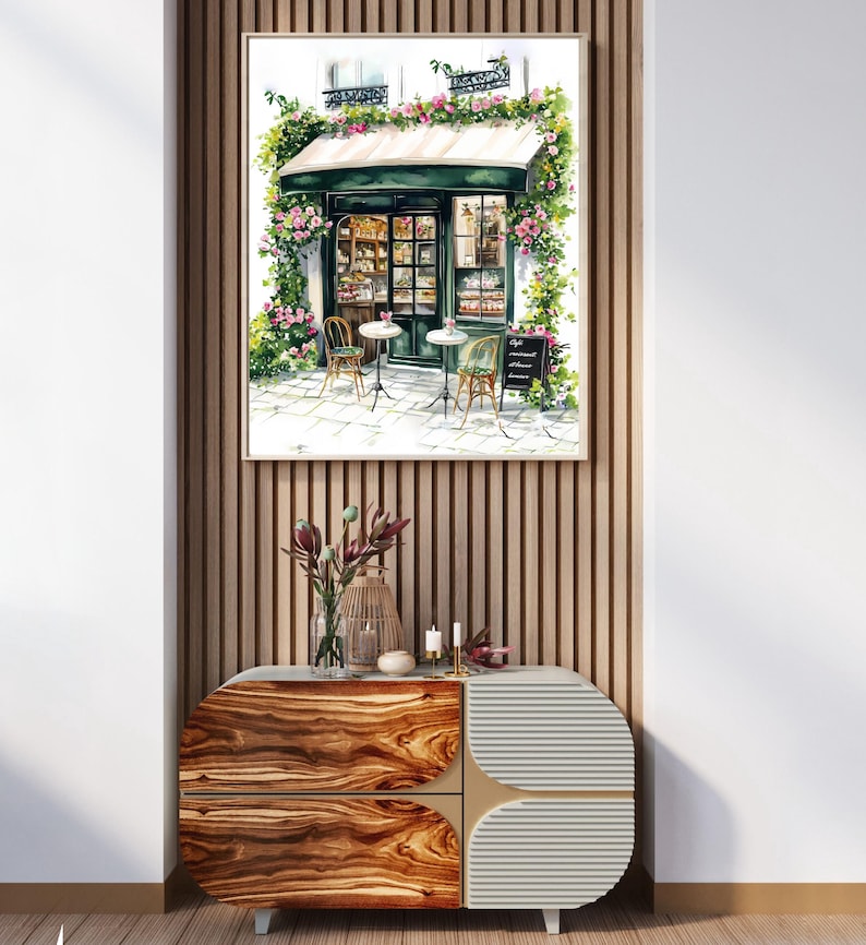 May include: A watercolor painting of a Parisian cafe with a green awning, flowers, and two tables with chairs. The cafe is surrounded by a white building with a window and a sign that reads "Cafe", "Gourmandises", and "Ap&eacute;ros".