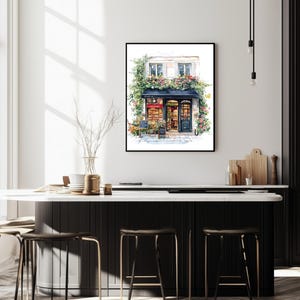 May include: A watercolor painting of a storefront with a black awning and a sign that reads "Le Petit March&eacute;". The storefront is decorated with flowers and greenery. The building is white with black trim. The painting is framed in a black frame.