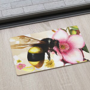 May include: A doormat with a bee and pink flowers. The bee is yellow and black with brown wings. The flowers are pink with white centers.