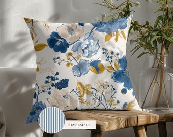 May include: A square throw pillow featuring a white background with a floral pattern in shades of blue, cream, and mustard yellow. A circular inset shows the reverse side of the pillow, which has a blue and white vertical striped pattern.