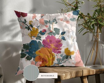 May include: A square throw pillow featuring a white background with a painterly floral pattern in shades of magenta, mustard yellow, teal, and soft pink. A circular inset shows a black and white vertical striped pattern labeled with the text REVERSIBLE.