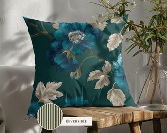 May include: A square throw pillow featuring a dark teal background with large, detailed blue flowers and light gray leaves. A circular inset shows the reverse side of the pillow, which has a pattern of thin, vertical, light and dark gray stripes.