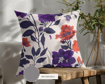 May include: A square throw pillow featuring a floral pattern with deep purple and bright orange flowers on a cream background. A circular inset shows the reverse side of the pillow, which has a black and white vertical stripe pattern with the text REVERSIBLE.