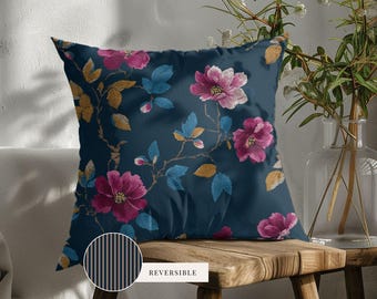 May include: A square throw pillow featuring a dark blue background with a floral pattern of pink flowers, blue leaves, and gold foliage. A circular inset shows the reverse side, which has a pattern of thin vertical black and tan stripes.