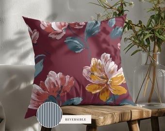 May include: A square throw pillow featuring a deep burgundy background with a floral pattern of large pink, yellow, and teal flowers and leaves. A small circular inset shows the reverse side, which has a dark blue and white vertical stripe pattern.