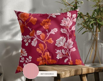 May include: A square throw pillow featuring a deep magenta background with a repeating floral pattern in shades of orange and white. A circular inset shows the reverse side of the pillow, which has a pattern of thin vertical white and pink stripes.