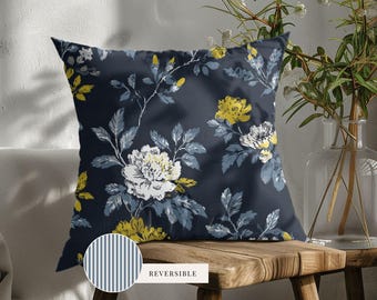 May include: A square throw pillow featuring a dark navy blue background with a floral pattern of white and yellow flowers and grey leaves. A circular inset shows the reverse side of the pillow, which has a blue and white vertical stripe pattern.