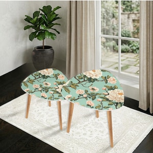 May include: Two matching side tables with a floral design. The tables have a light blue background with white and peach-colored flowers and green leaves. The legs are made of light-colored wood.