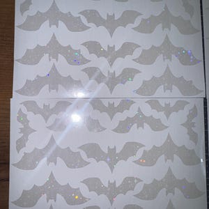 Puede incluir: Hojas de pegatinas blancas con forma de murciélago con un efecto brillante e iridiscente. Las pegatinas están dispuestas en filas y columnas, perfectas para manualidades y decoraciones de Halloween. Los murciélagos tienen un diseño de alas detallado.