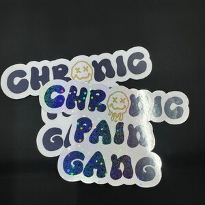 May include: Two holographic stickers with the text "Chronic Pain Gang" in a blue and white glittery font. The stickers feature a yellow smiley face with an "X" for eyes.