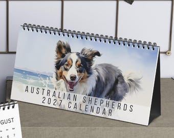 May include: A desk calendar featuring Australian Shepherds with a spiral binding. One page displays a watercolor-style image of a dog in a forest with the month of August, while the other shows a dog on a beach with the text Australian Shepherds 2027 Calendar.