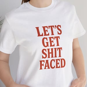 May include: White t-shirt with the phrase "LET'S GET SHIT FACED" printed in red. The shirt is a crew neck style and is being worn by a person. The person is wearing blue jeans.