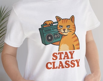 May include: White t-shirt featuring an orange tabby cat holding a teal boombox. The cat has a stern expression. The words "STAY CLASSY" are printed in orange below the cat. The shirt is made of a soft material.