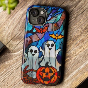 May include: A colorful phone case featuring a Halloween-themed stained glass design. The case showcases two smiling ghosts, a jack-o'-lantern, and bats against a blue and purple background. The case is on a wooden surface.