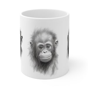 Peut inclure: Une tasse en céramique blanche avec une illustration en noir et blanc du visage d'un bébé singe. Le singe sourit et a un visage duveteux et poilu.