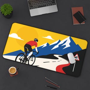May include: A rectangular mousepad featuring a cyclist riding uphill on a road with a mountain backdrop. The design includes a yellow sky with a sun and clouds, and a road sign indicating a 10% grade.