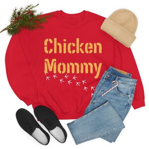 May include: Red sweatshirt with the text "Chicken Mommy" in yellow with white chicken footprints below. The sweatshirt is laid out on a white surface with a pair of black shoes, blue jeans, and a beige beanie.