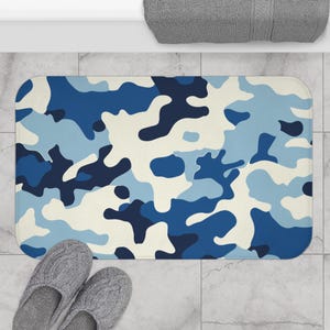 Può includere: Un tappetino da bagno rettangolare con un motivo mimetico blu in varie tonalità di blu e bianco sporco. Il tappetino è posizionato su un pavimento piastrellato, con un paio di pantofole grigie nella parte inferiore. Il motivo ricorda il camouflage militare.