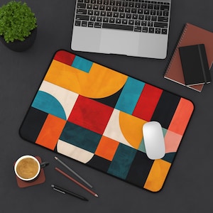 May include: A rectangular mouse pad with a colorful geometric design. The pattern features orange, yellow, red, blue, and black shapes. A white computer mouse sits on the pad, with a laptop and other office supplies in the background.