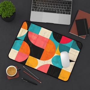 May include: A colorful geometric design mousepad with a white computer mouse on top. The design features circles, squares, and rectangles in shades of orange, yellow, blue, pink, and black.