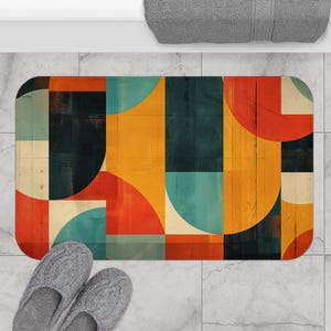 May include: A rectangular bath mat with a colorful geometric design. The pattern features orange, teal, and black shapes. The mat is placed on a light gray tiled floor. A pair of gray knit slippers are at the bottom of the mat.
