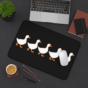 May include: Black rectangular mousepad featuring a line of five white cartoon ducks with orange beaks and feet. A white computer mouse rests on the mousepad. A laptop, notebooks, and a cup of coffee are also visible.