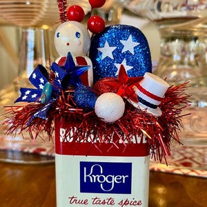 May include: A patriotic decorative arrangement in a vintage Kroger spice tin. The arrangement includes a wooden doll, glittery stars, red and blue accents, and a miniature top hat. The tin reads "true taste spice" and "NET WT. ½ OZ."