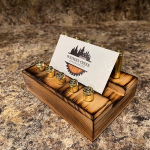 May include: A wooden business card holder with a burnt finish. The holder has slots for business cards and is decorated with spent bullet casings. The business card in the holder reads "Whiskey Creek Woodworking".
