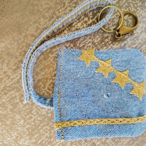 May include: A light blue denim key card holder with a gold trim and three gold star embellishments. The holder has a matching denim strap and a gold-colored metal clasp.