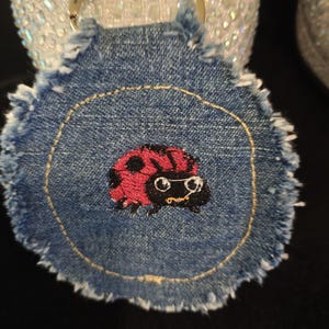 Peut inclure: Un porte-clés circulaire en jean avec un bord effiloché et un motif central brodé représentant une coccinelle rouge à pois noirs portant de petites lunettes blanches. Le tissu en jean est bleu moyen avec une couture circulaire jaune.