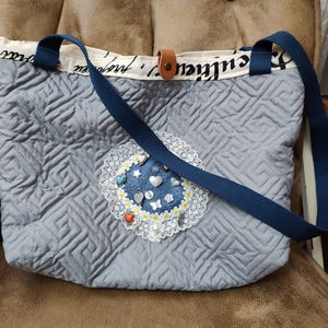 May include: A quilted gray tote bag with a blue strap and decorative embellishments. The bag features a denim patch adorned with heart and butterfly charms, surrounded by a lace trim. The bag has a cream-colored top with black script.