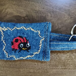 May include: Blue denim keychain featuring an embroidered ladybird design. The ladybird is red with black spots and a yellow face. The keychain has a silver clasp and a denim strap. The ladybird is stitched on a blue denim background.