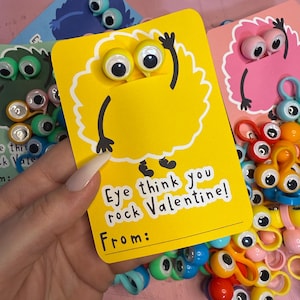 May include: A yellow Valentine's Day card with googly eyes and the text "Eye think you rock Valentine!" is held in a hand. The card has a cartoon monster design. Other cards and googly eye accessories are in the background.