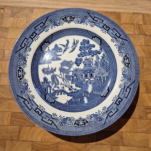 May include: A blue and white ceramic plate with a Willow pattern design. The plate features a central scene with a pagoda, bridge, and figures, surrounded by a decorative border. The plate is round and has a classic, vintage aesthetic.