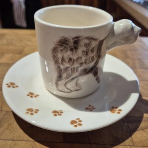 May include: A white ceramic teacup and saucer set. The cup has a brown illustration of a dog's fur and a dog-shaped handle. The saucer is decorated with brown paw prints. The set is designed for serving tea or coffee.