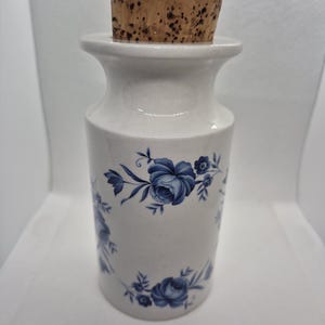 May include: White ceramic jar with a cork stopper. The jar is decorated with blue floral patterns. The jar has a cylindrical shape with a wider base and a narrower neck. The cork stopper is a light brown colour.