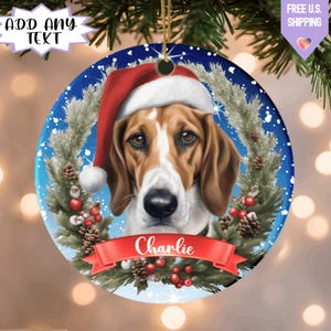 May include: Round Christmas ornament featuring a dog wearing a Santa hat. The dog has brown and white fur and is surrounded by a wreath of pine needles, berries, and pine cones. The name "Charlie" is displayed on a red banner.