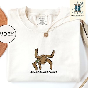 May include: Ivory-colored t-shirt with an embroidered brown and green robot design. The text "AMAZE AMAZE AMAZE" is below the robot. The shirt has a crew neck and short sleeves. A gold necklace with a pendant is visible.