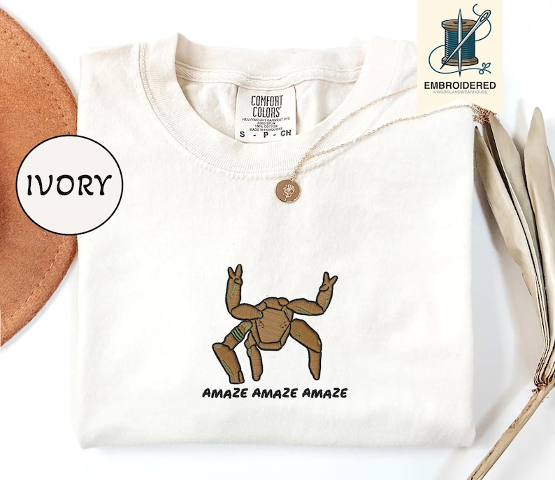 May include: Ivory-colored t-shirt with an embroidered brown and green robot design. The text "AMAZE AMAZE AMAZE" is below the robot. The shirt has a crew neck and short sleeves. A gold necklace with a pendant is visible.