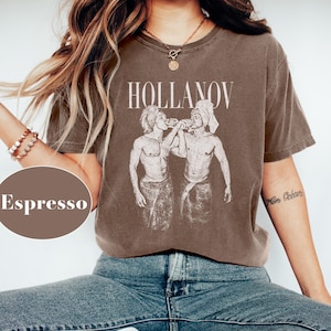 May include: Espresso-colored t-shirt with the word "HOLLANOV" in a serif font above a graphic of two figures. The figures are depicted in a vintage style, with one wearing a towel on their head. The shirt is paired with blue jeans.