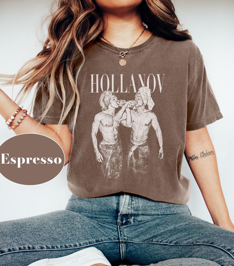 May include: Espresso-colored t-shirt with the word "HOLLANOV" in a serif font above a graphic of two figures. The figures are depicted in a vintage style, with one wearing a towel on their head. The shirt is paired with blue jeans.