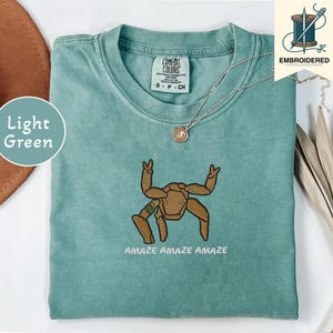 May include: Light green t-shirt with an embroidered brown crab design and the words "AMAZE AMAZE AMAZE" in white. The shirt has a crew neck and short sleeves. A silver necklace with a small pendant is visible.